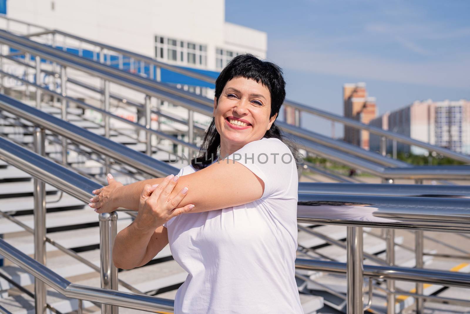 Smiling senior woman doing stretching outdoors on urban background by Desperada