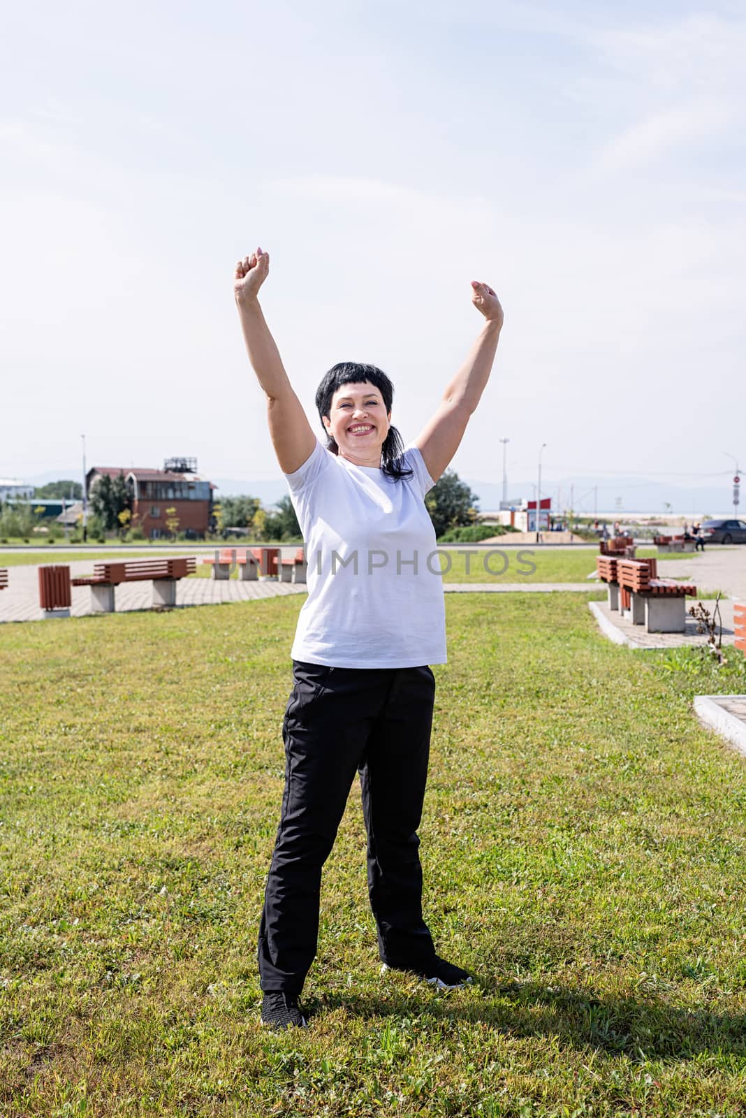 Happy senior woman in sports clothes exercising in the park standing with arms up by Desperada