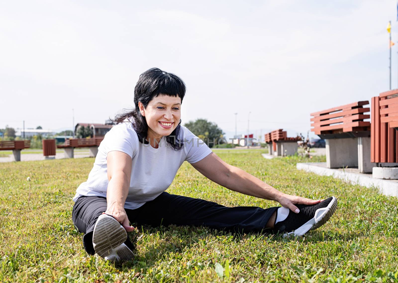 Smiling senior woman warming up stretching sitting on the grass in the park by Desperada