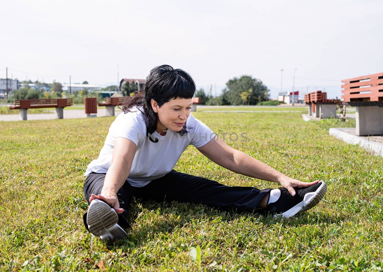 Smiling senior woman warming up stretching sitting on the grass in the park by Desperada