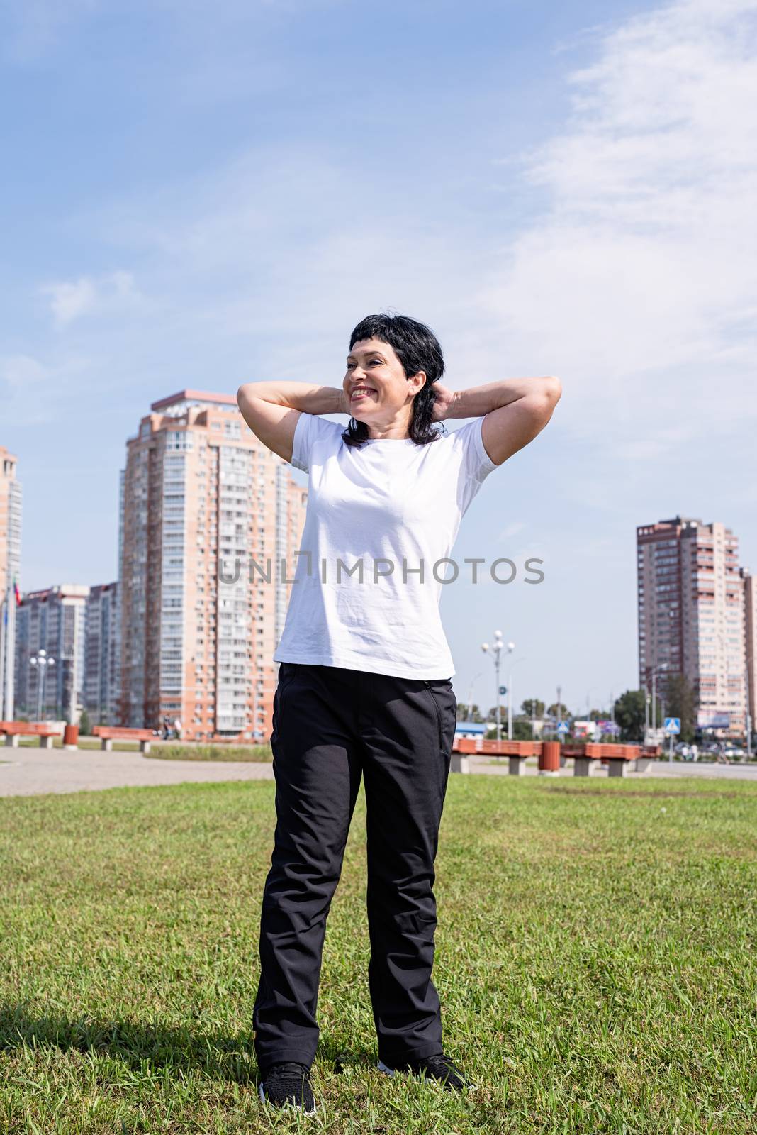 Smiling senior woman warming up before training outdoors in the park on urban background by Desperada