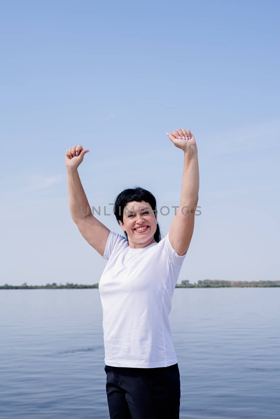 Sport and fitness. Senior sport. Active seniors. Active and happy senior woman exercising near the riverside standing with arms up