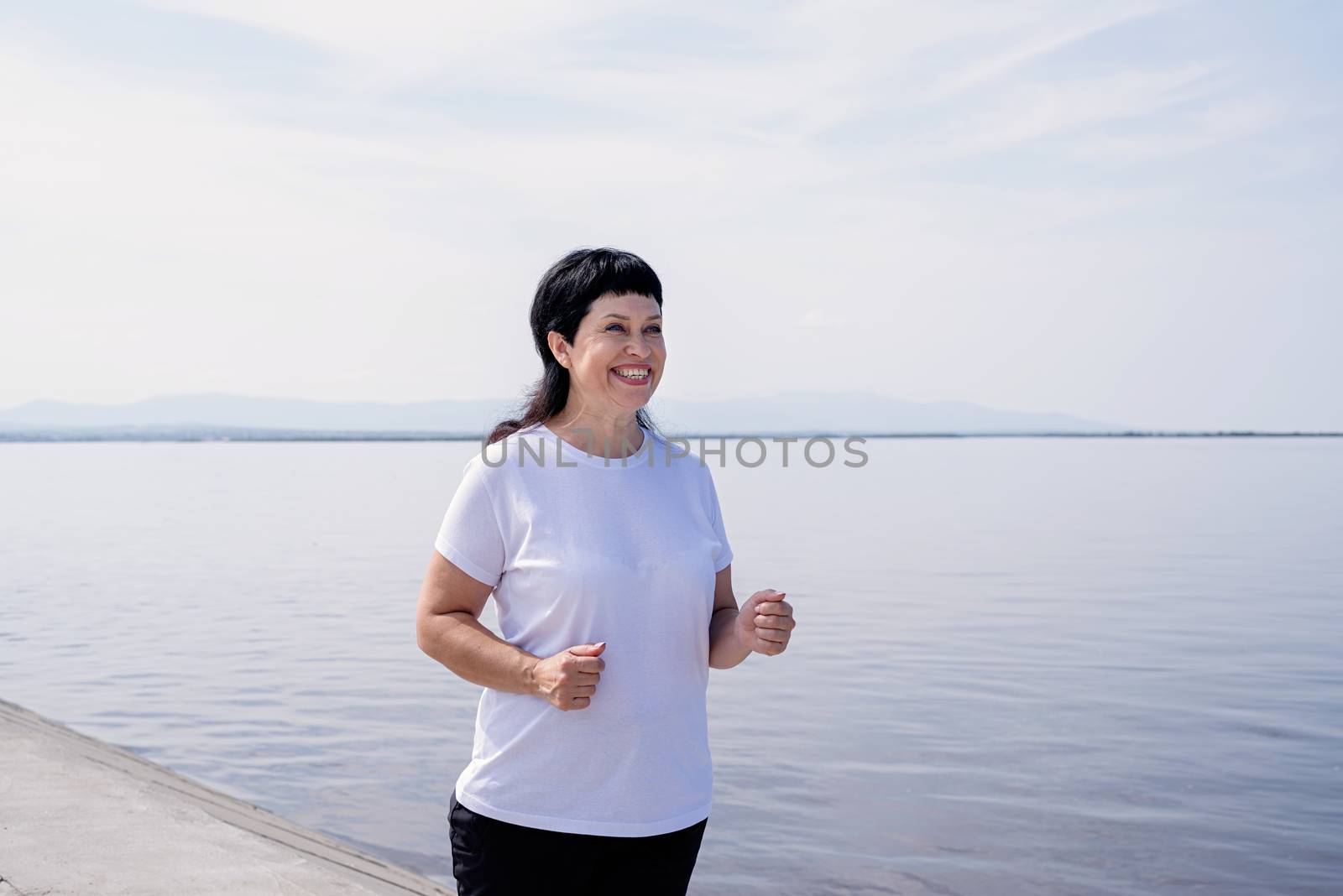 Active laughing senior woman jogging near the riverside by Desperada
