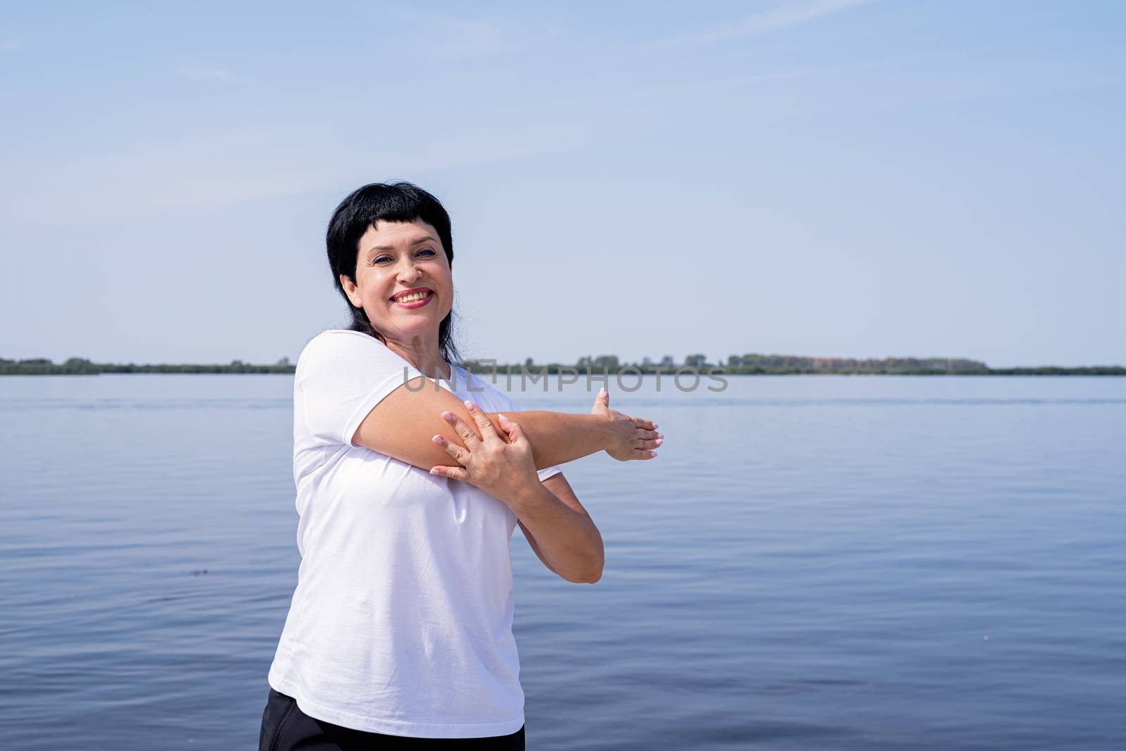 Active and happy senior woman doing stretching near the riverside by Desperada