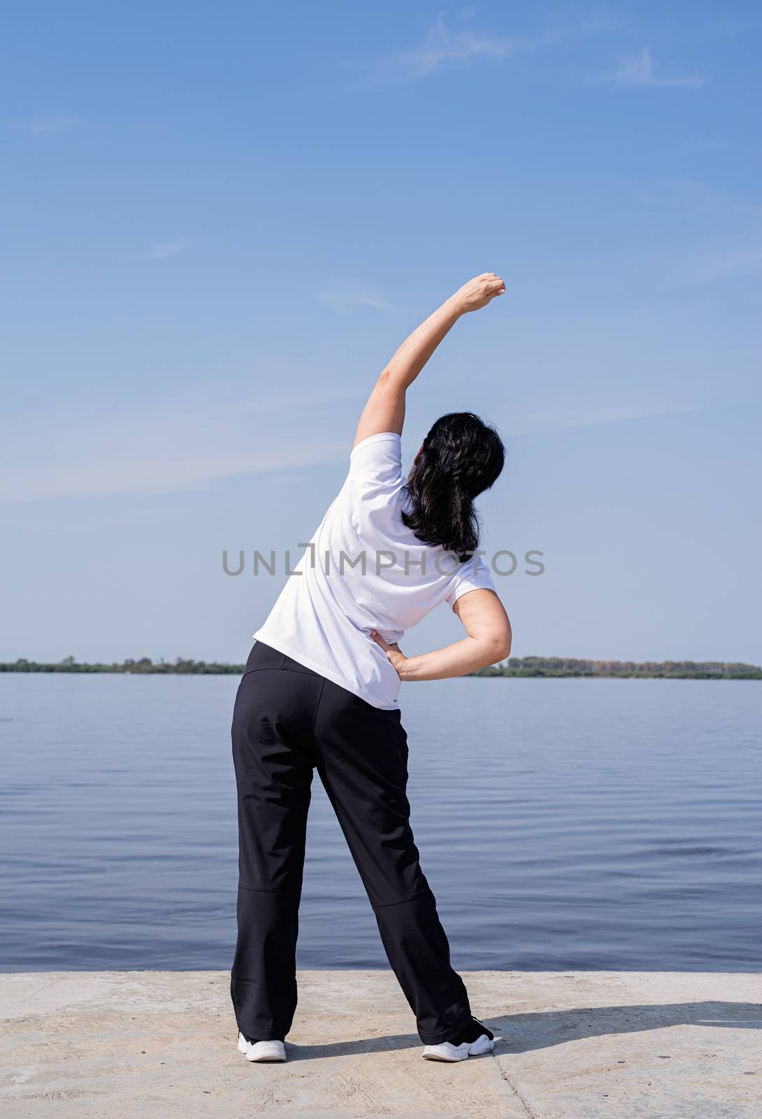 Active and happy senior woman doing stretching near the riverside by Desperada