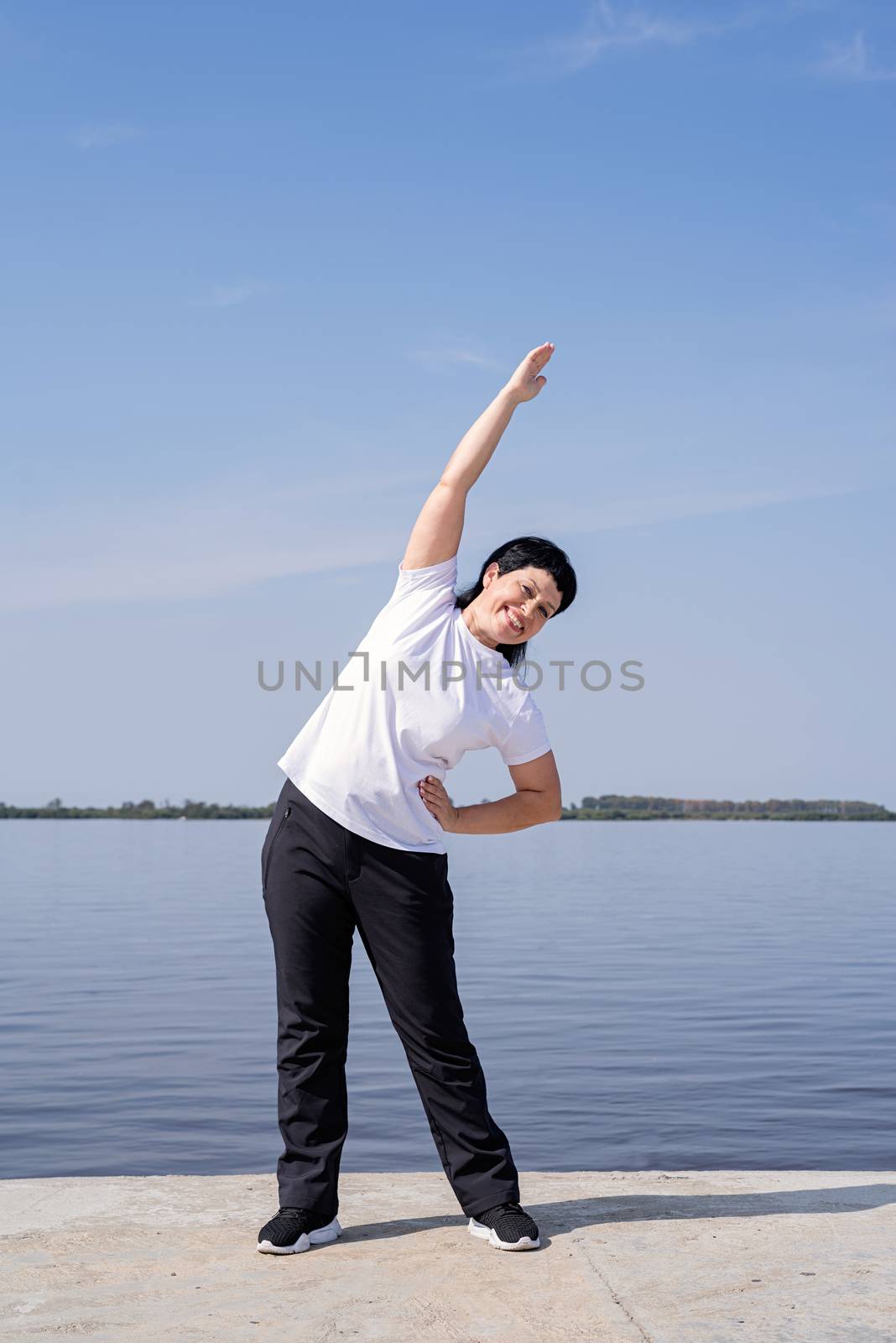 Active and happy senior woman doing stretching near the riverside by Desperada