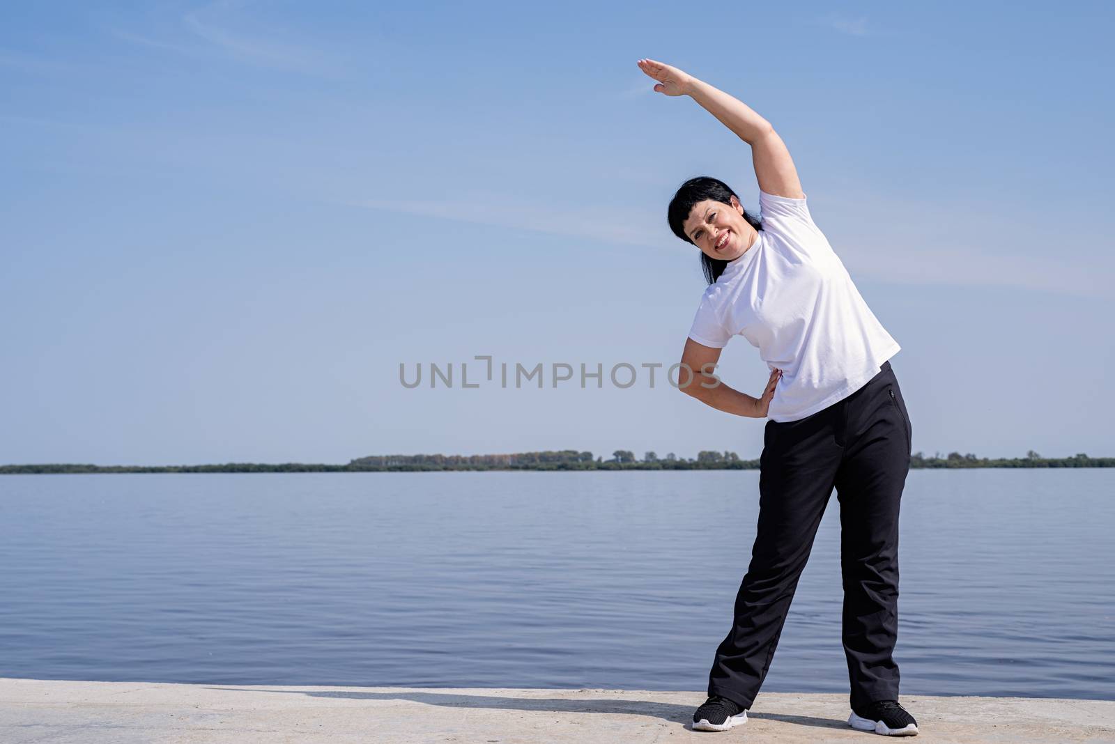 Active and happy senior woman doing stretching near the riverside by Desperada