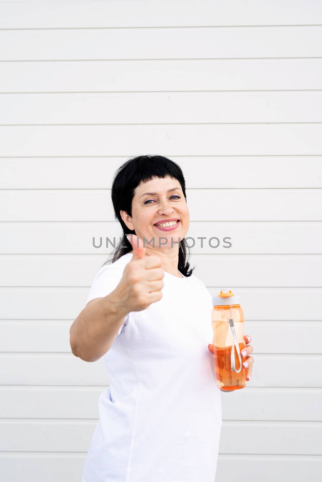 senior woman drinking water and showing thumbs up after workout outdoors on urban background by Desperada