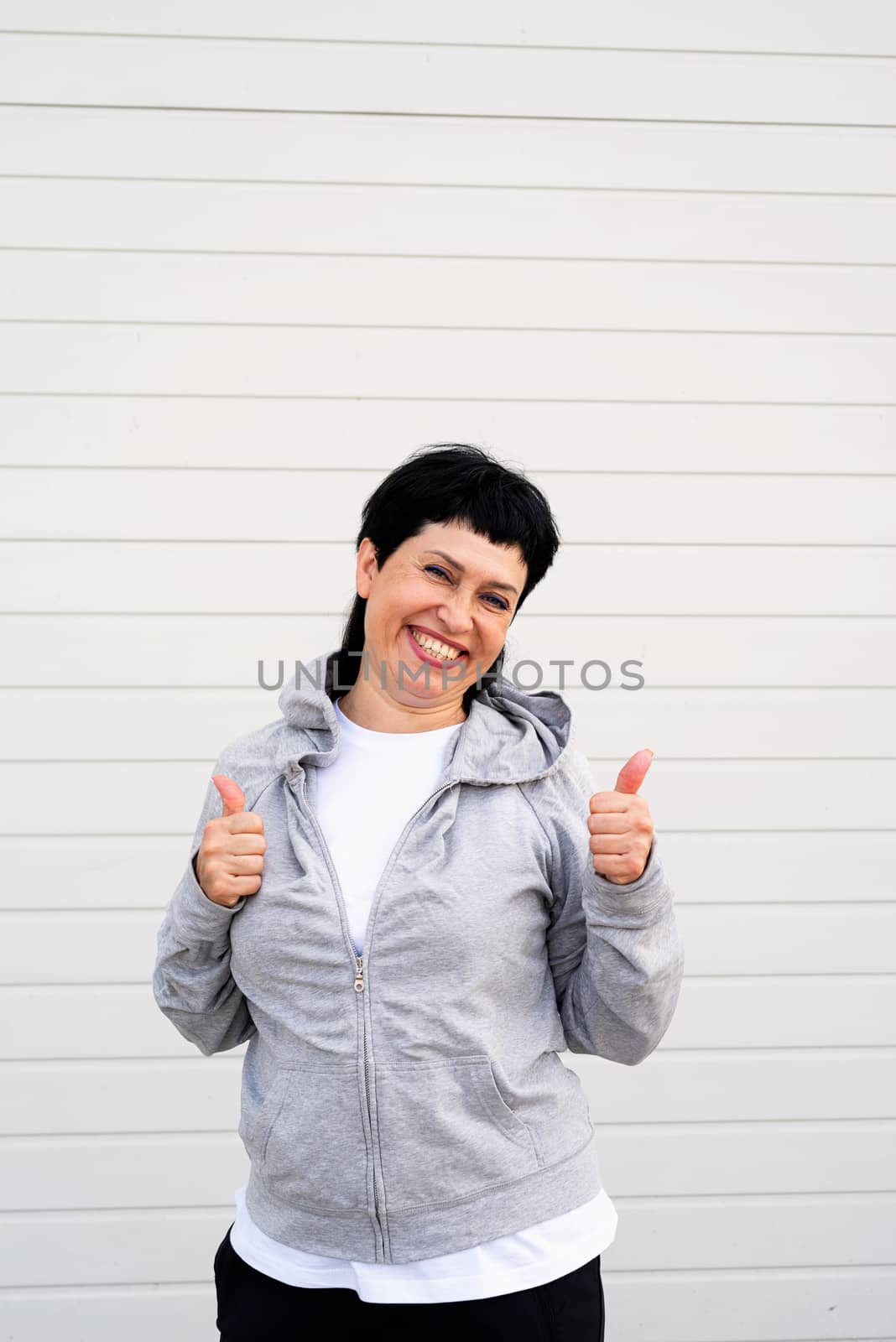 Sport and fitness. Senior sport. Active seniors. Senior woman showing thumbs up standing outdoors on gray solid background