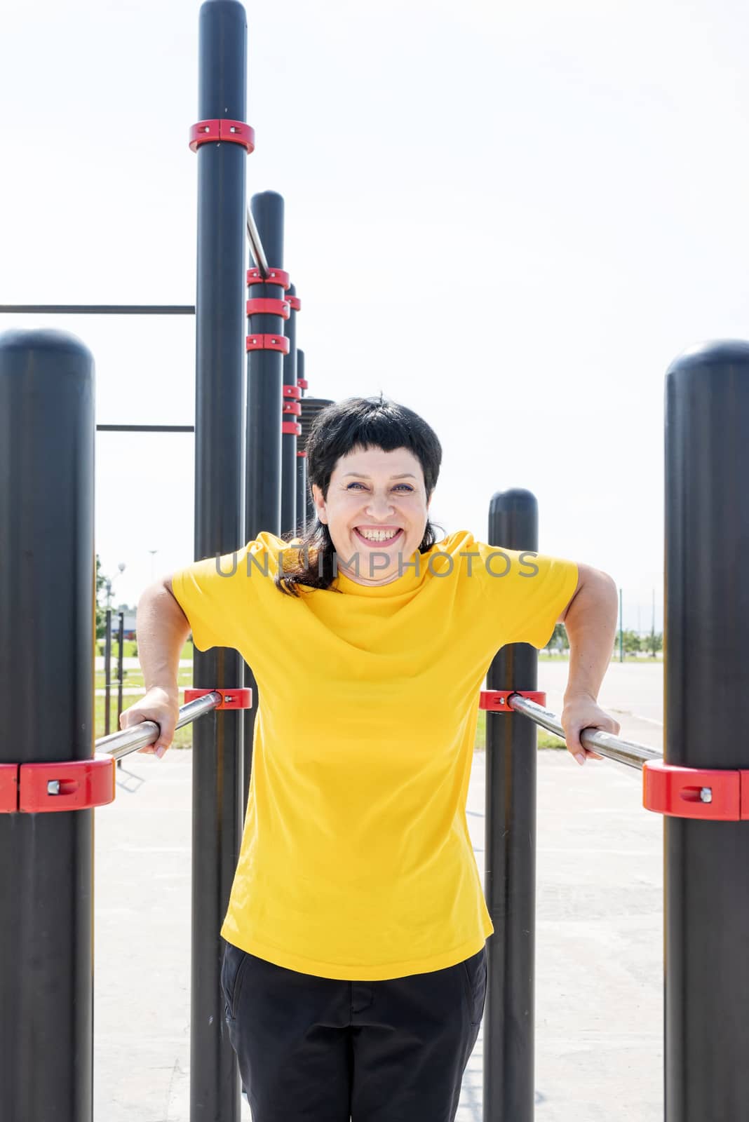 Smiling senior woman doing reverse push ups outdoors on the sports ground bars by Desperada