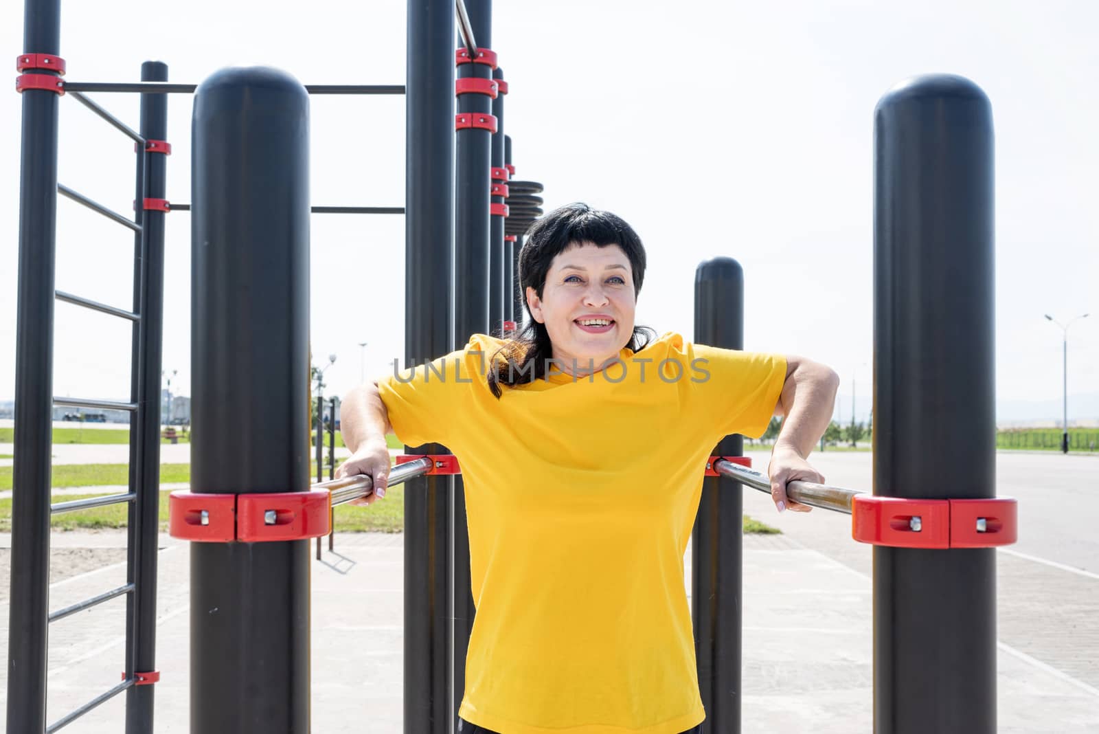 Smiling senior woman doing reverse push ups outdoors on the sports ground bars by Desperada