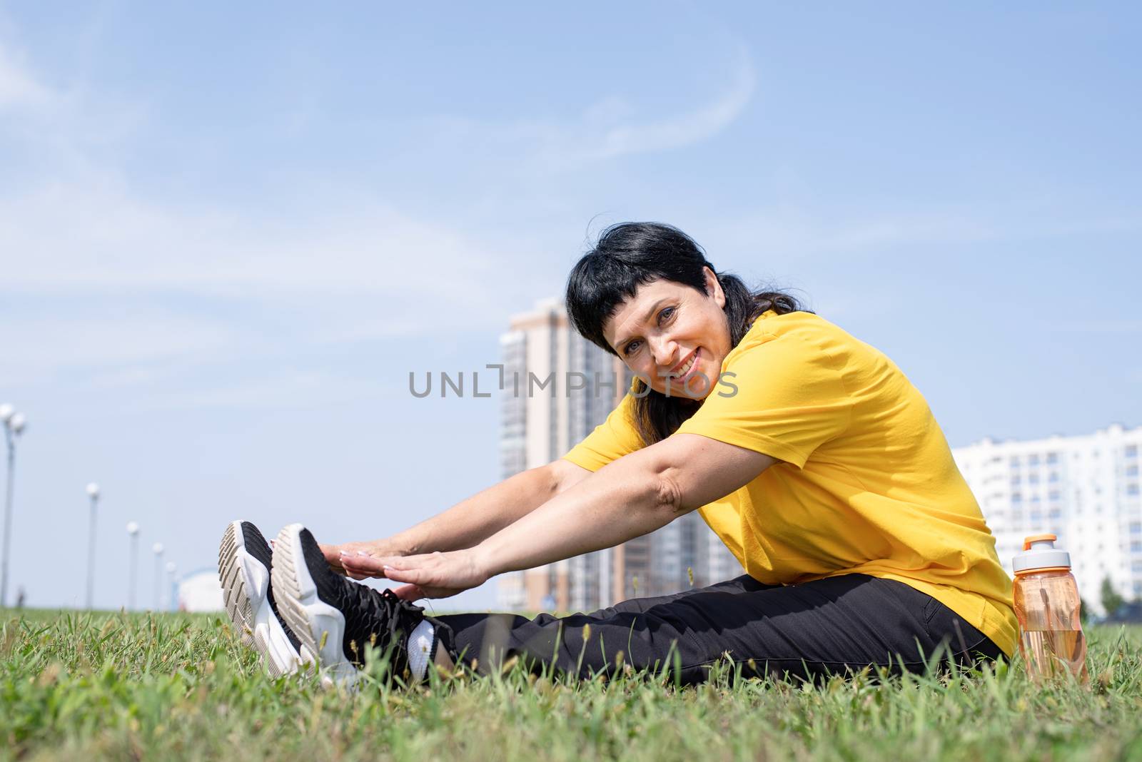 Senior woman stretching her legs on grass in the park on urban background by Desperada