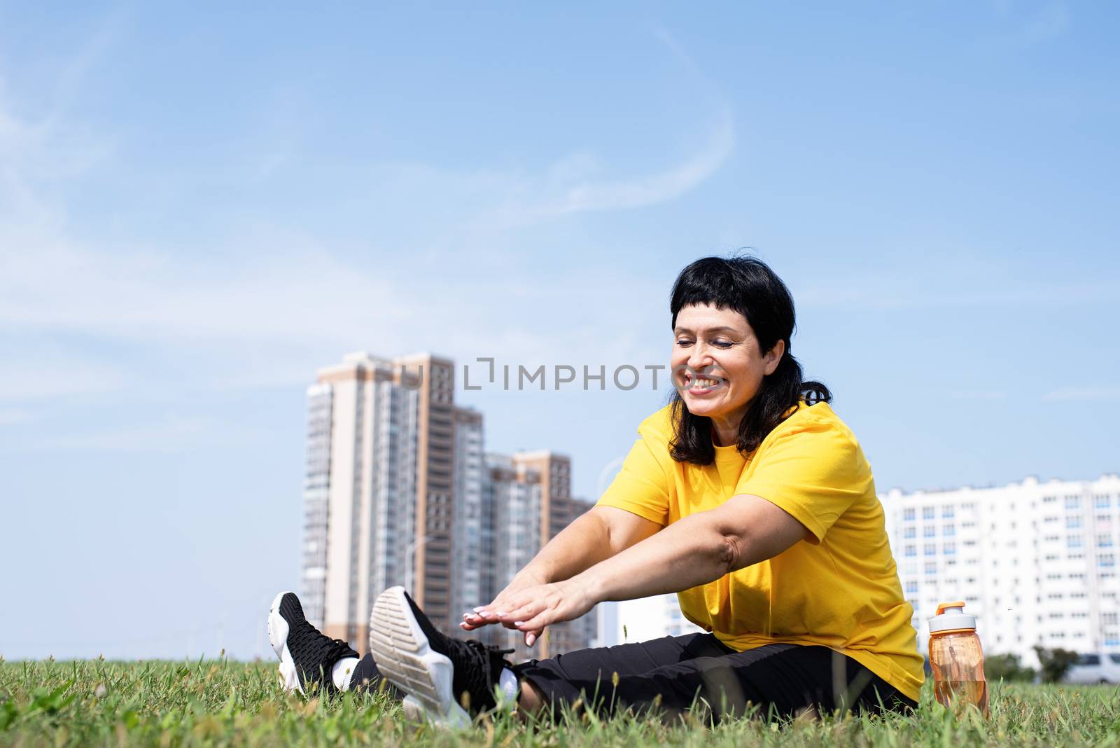 Sport and fitness. Senior sport. Active seniors. Senior woman stretching her legs on grass in the park on urban background