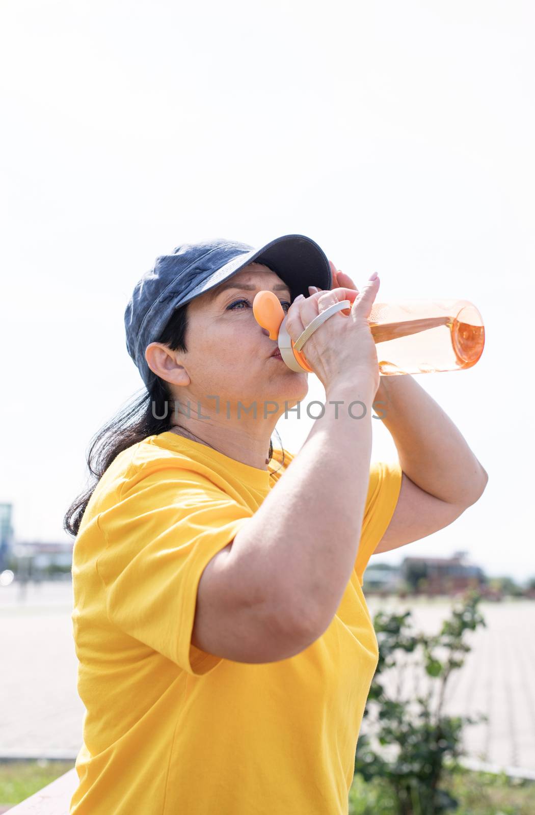 Smiling senior woman drinking water after workout outdoors on the sports ground by Desperada