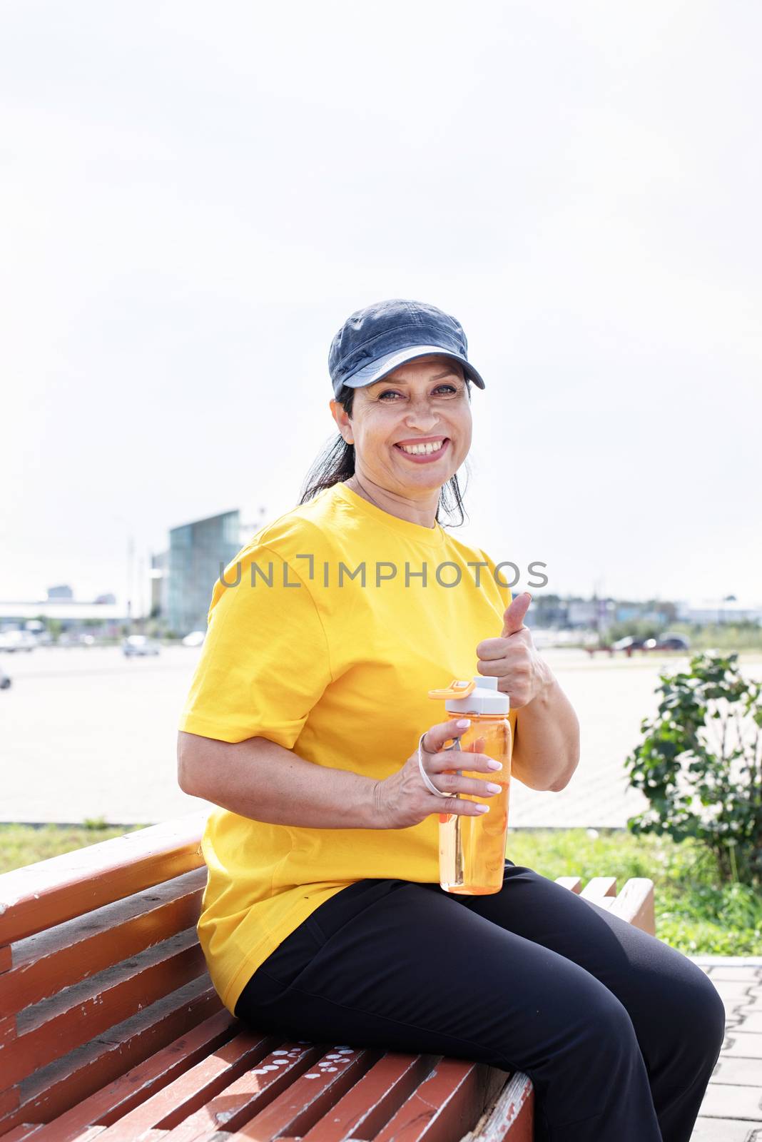 Sport and fitness. Senior sport. Active seniors. Smiling senior woman drinking water after workout outdoors on the sports ground showing thumbs up