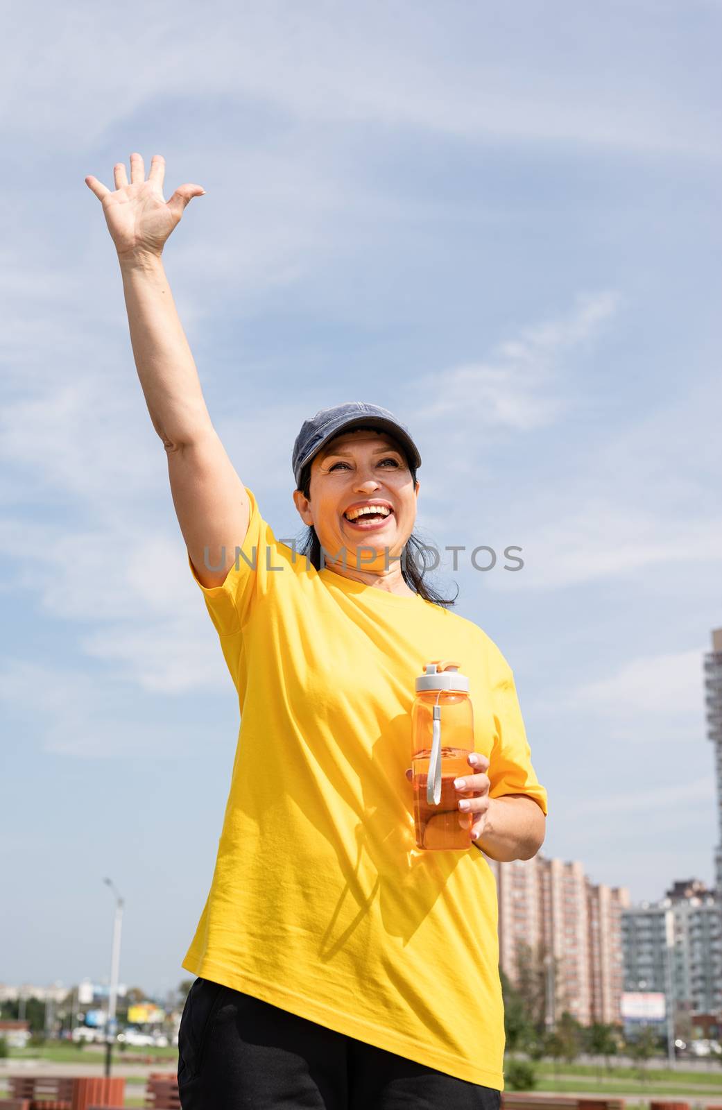 Excited senior woman saying hello drinking water after workout outdoors on the sports ground by Desperada