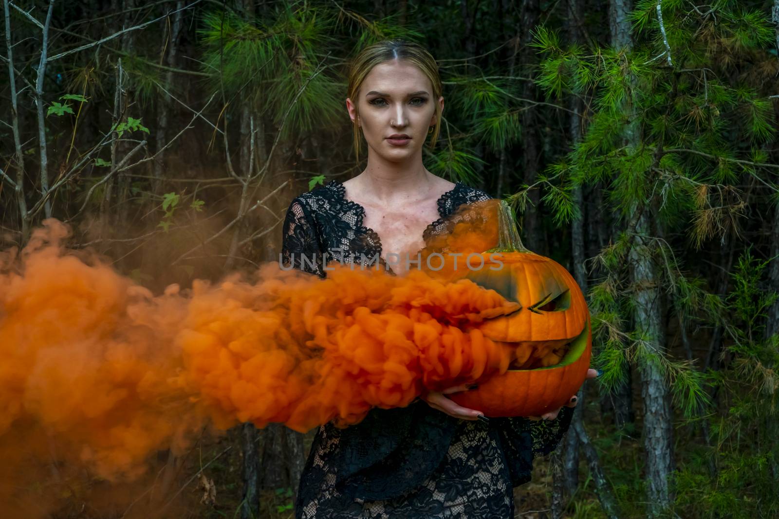 A beautiful blonde model wearing all black poses with a smoking pumpkin in an outdoor environment