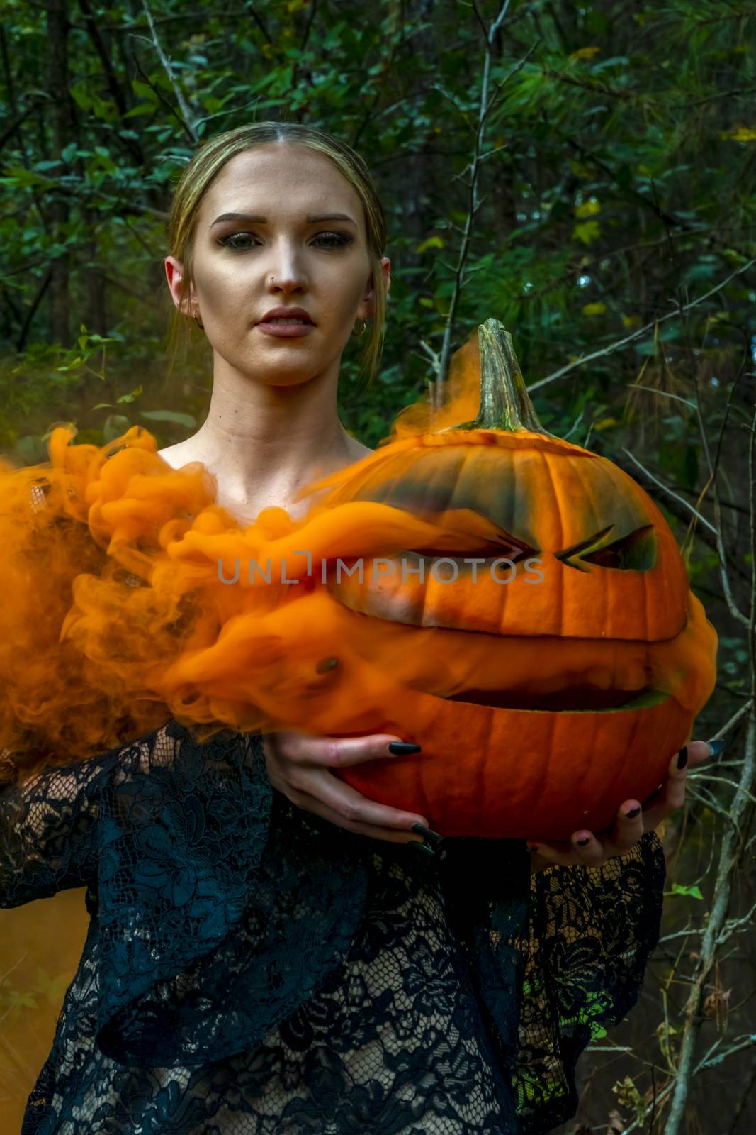 A beautiful blonde model wearing all black poses with a smoking pumpkin in an outdoor environment