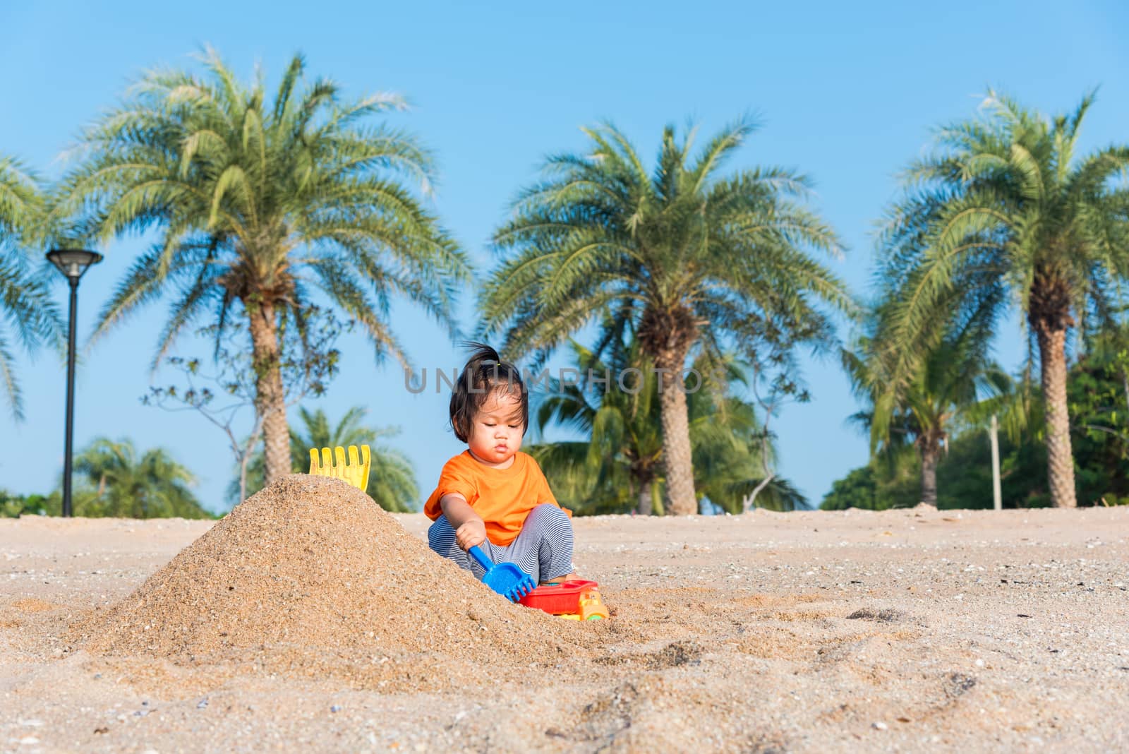 Asian Thai happy cute little cheerful daughter girl funny digging playing toy with sand at an outdoor tropical beach in summer day with copy space