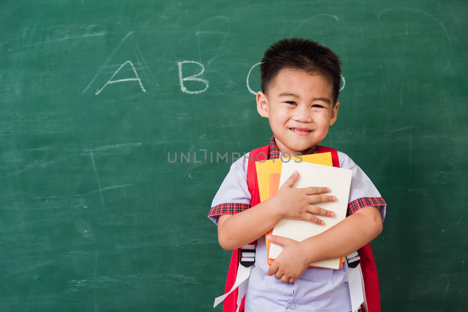 Back to School. Happy Asian funny cute little child boy from kindergarten in student uniform with school bag hold or hug books smile on green school blackboard, First time to school education concept