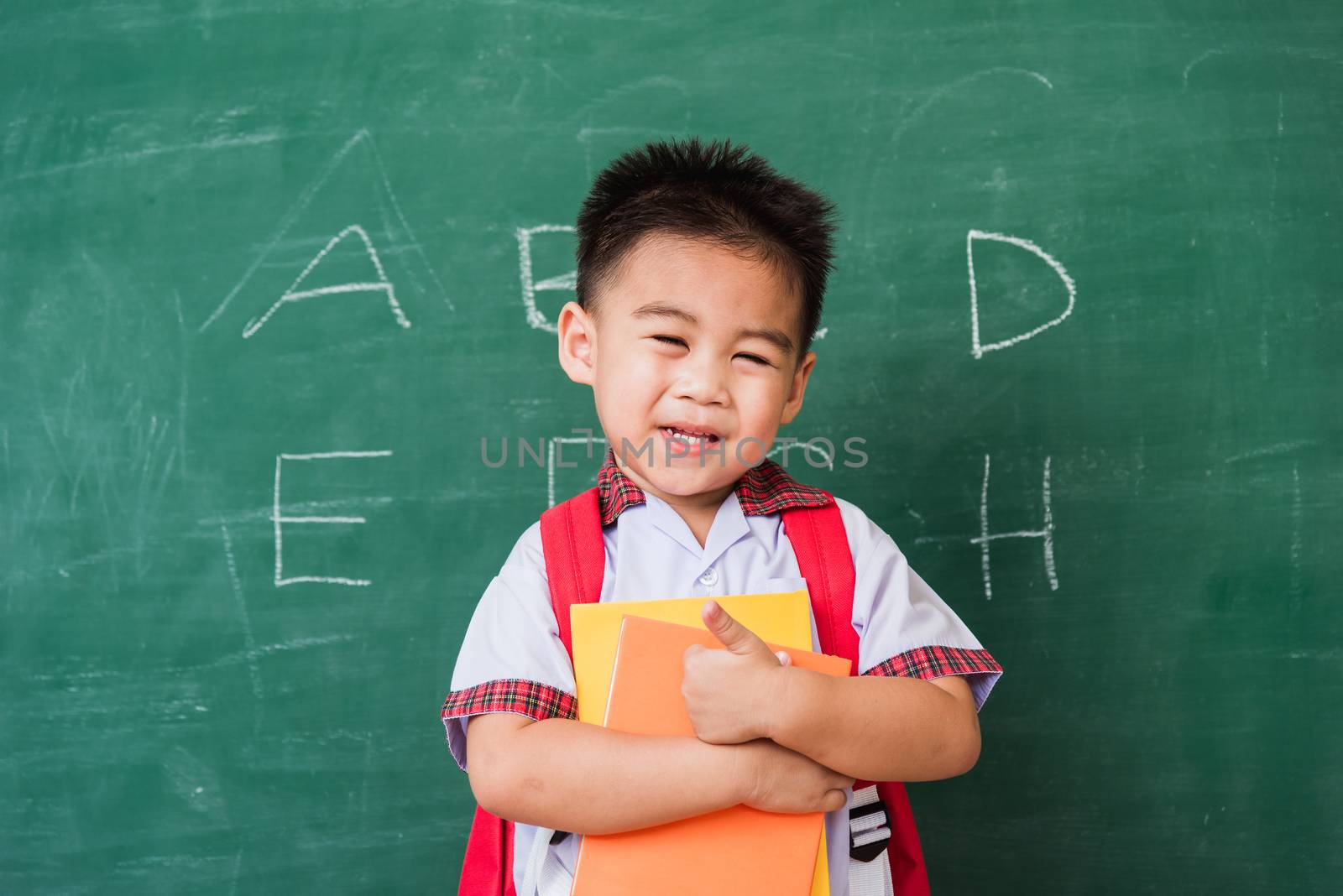 Back to School. Happy Asian funny cute little child boy kindergarten in student uniform with school bag and books smile show finger thumb up on green school blackboard, First time to school education