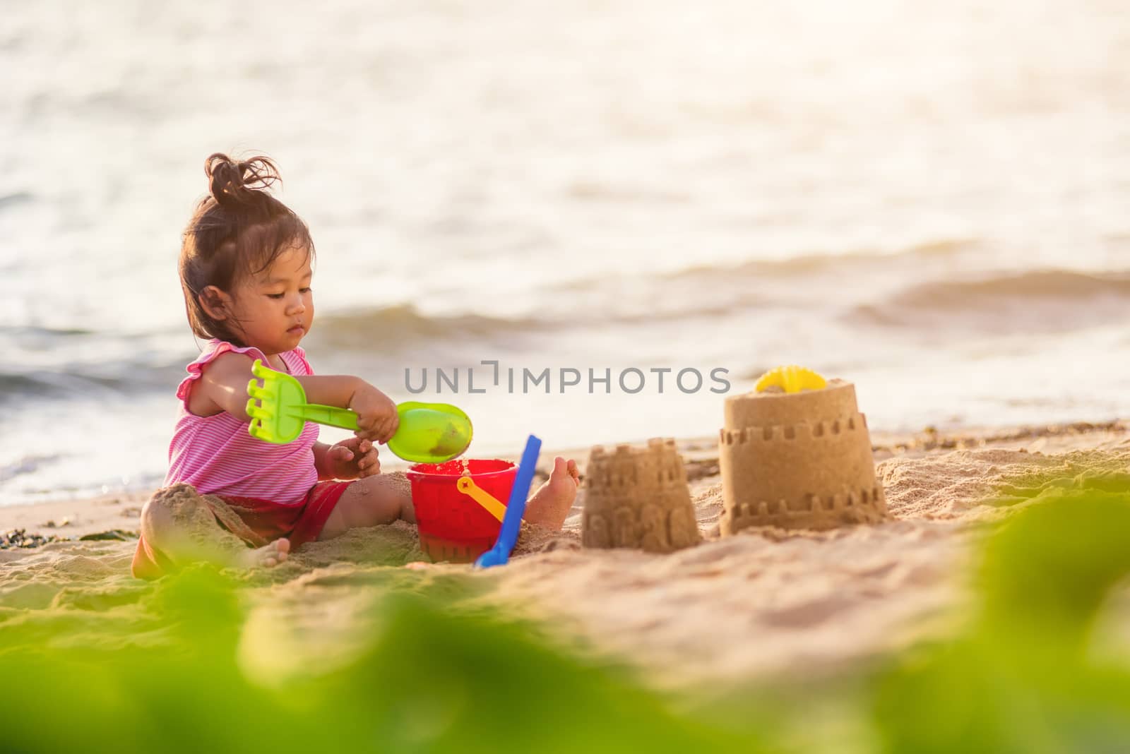 Happy fun Asian child cute little girl playing sand with toy sand tools at a tropical sea beach in holiday summer on sunset time, tourist trip concept