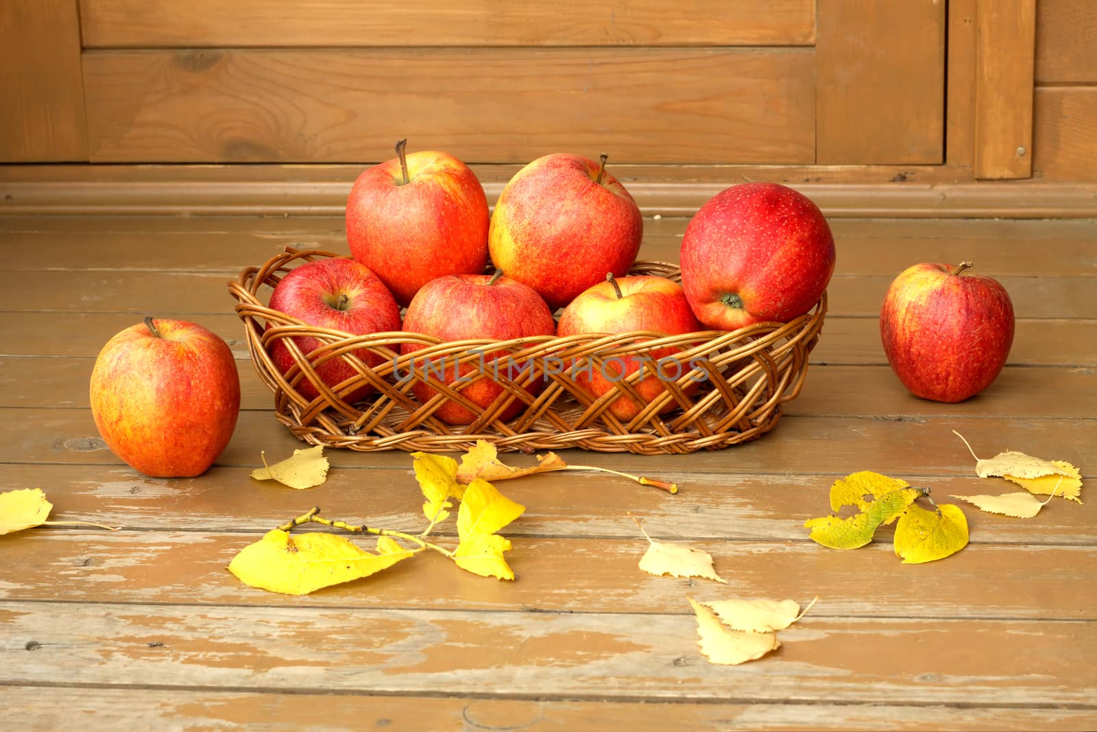 Autumn still life with ripe red apples in a wicker basket and yellow leaves scattered on a wooden surface closeup by dymaxfoto