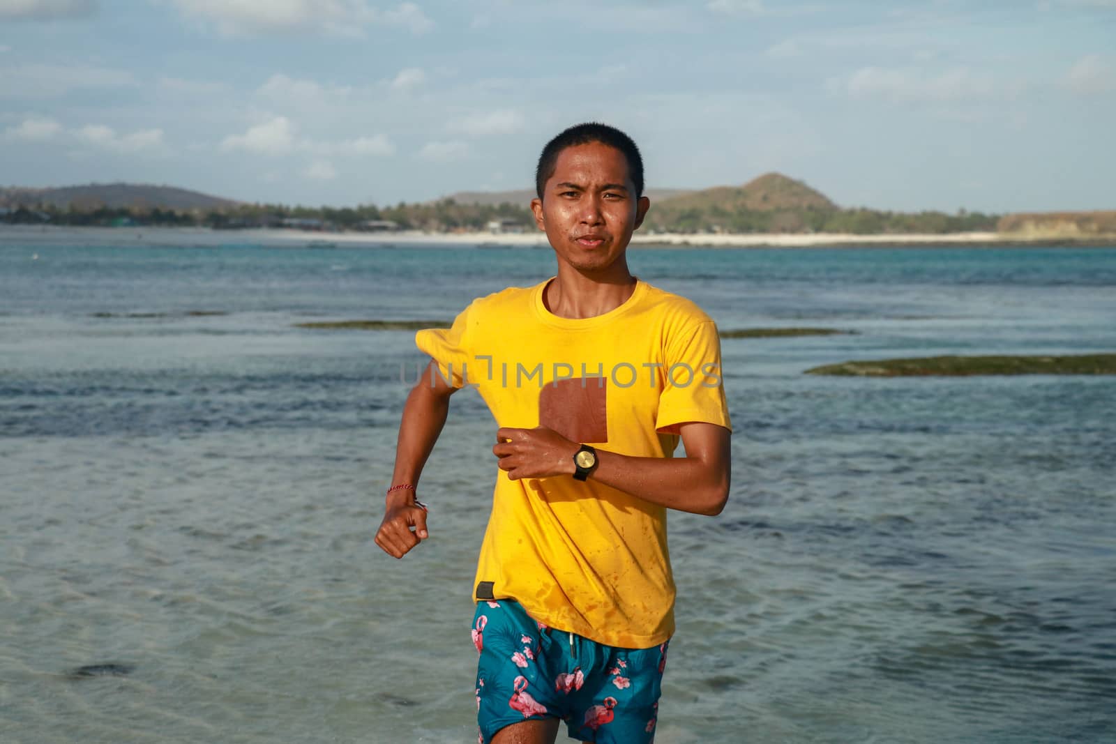 Man training on the beach in morning. Young man on morning run outdoors. Young asian in yellow t-shirt running along the beach. Teenager trains running by the sea.