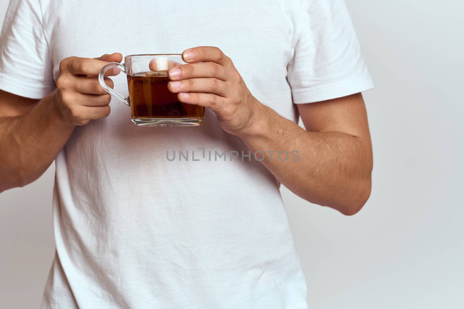 A man with a hot drink of tea in his hands in a white T-shirt on a light background cropped view. High quality photo