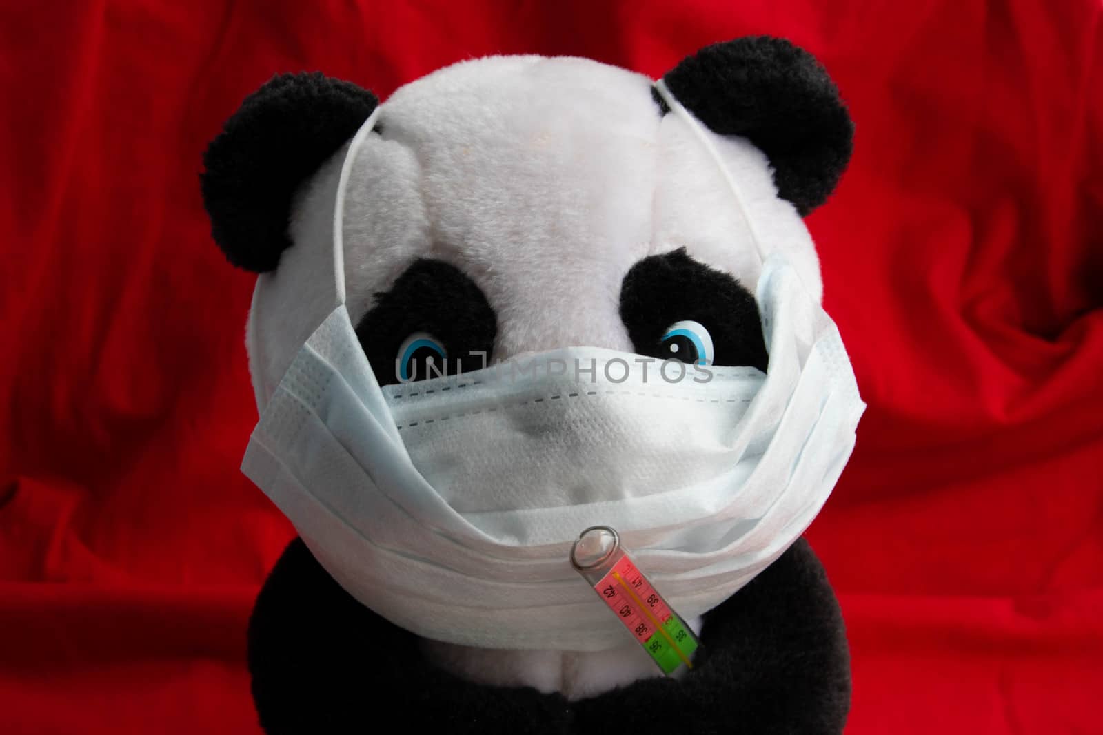 A toy panda in a medical mask with a thermometer sits on a red background. Coronavirus treatment concept