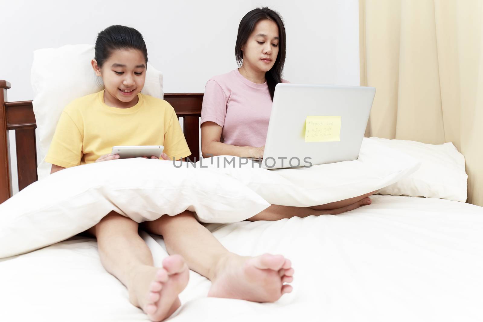 Asian mother and daugther looking laptop and tablet on bed in bedroom. Business women woking with laptop at home with her daugther smiling enjoy with tablet on bed. Work at home concept.