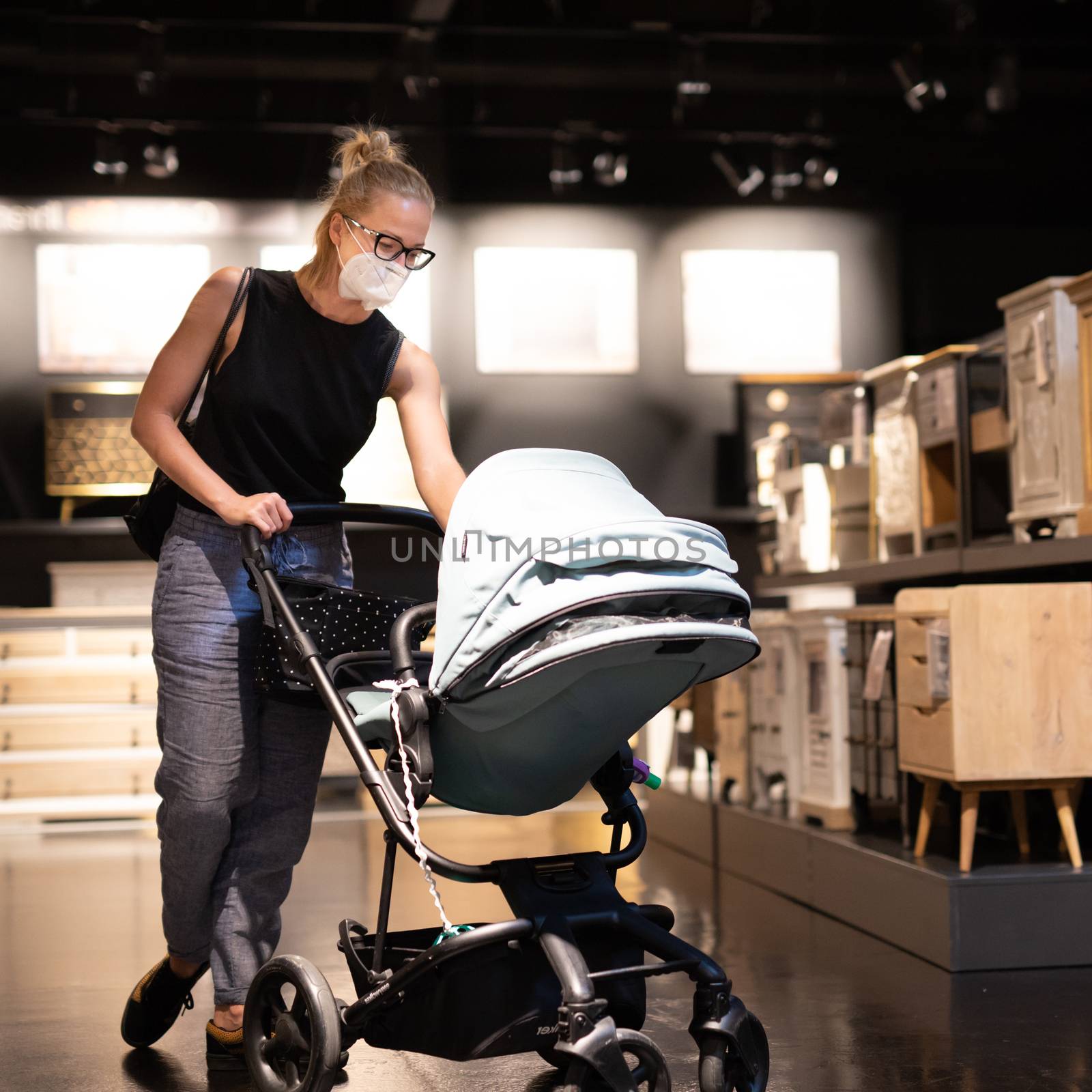 Young mom with newborn in stroller shopping at retail furniture and home accessories store wearing protective medical face mask to prevent spreading of corona virus. New normal during covid epidemic.