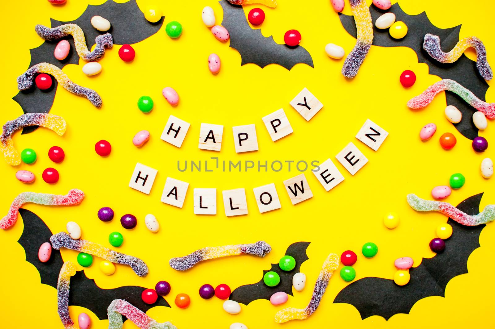inscription from wooden blocks Happy Halloween and frame made of paper homemade bats and multicolored candies and worms from gummy on a bright yellow background