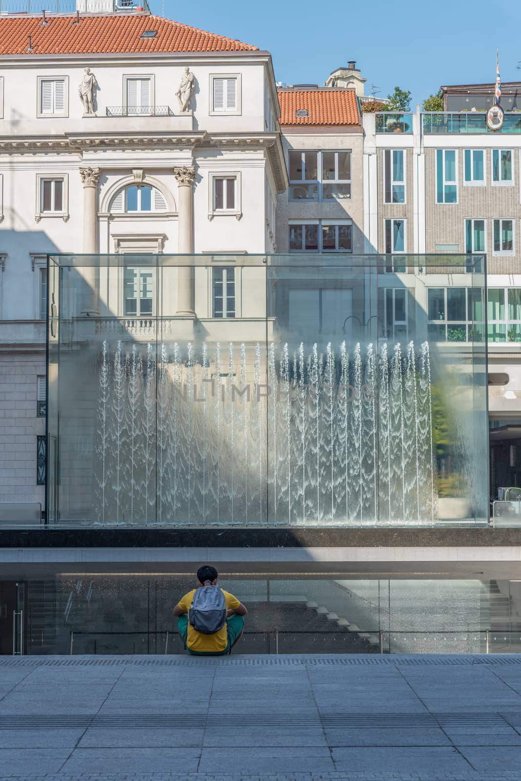 Person sitting back to back in front of a modern fountain splashing water on a huge glass window cut by the shadow, contemporary architecture in the city center