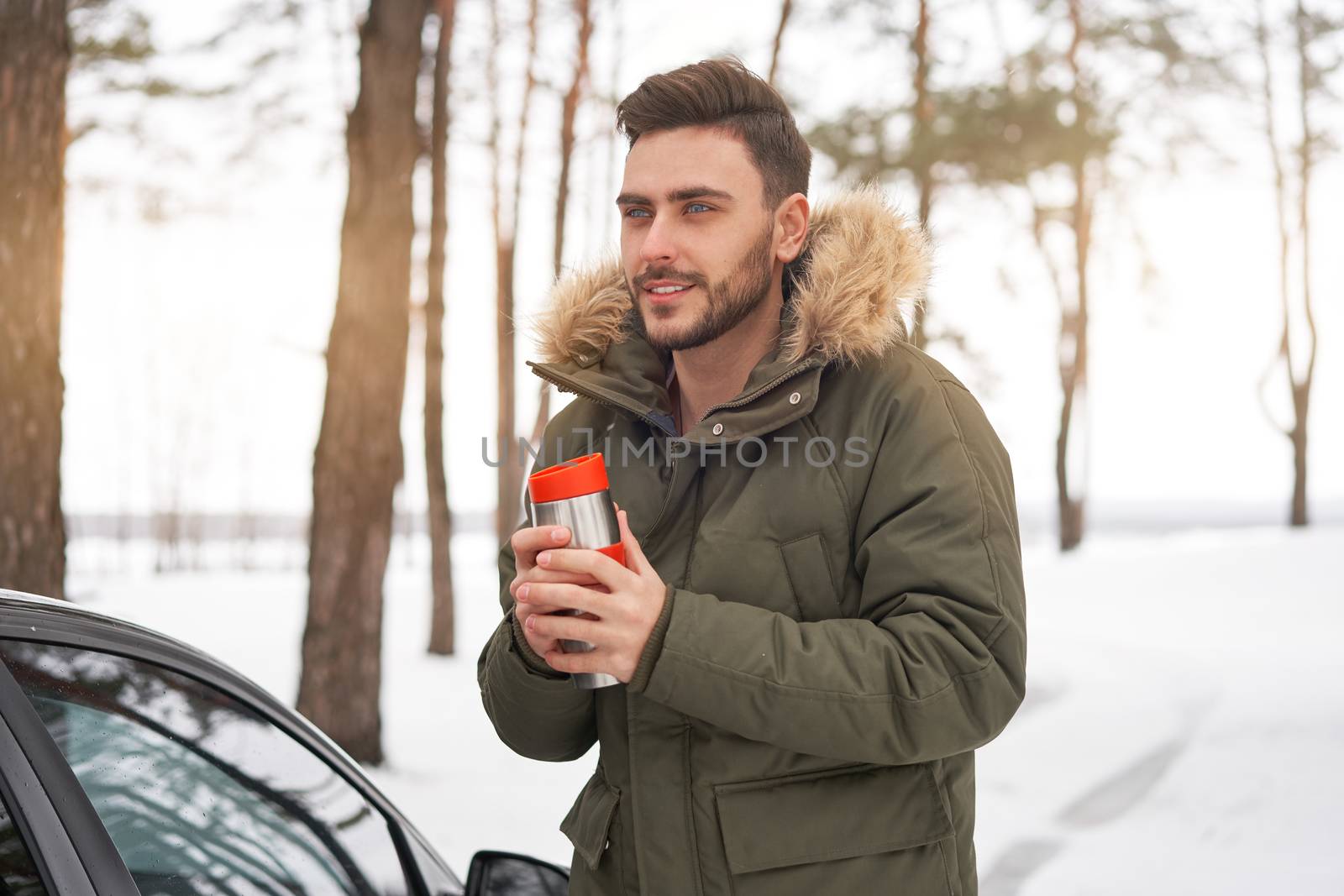 Attractive Caucasian man stands winter forest near car drink hot beverage thermos. Winter holiday road trip. Cold season. Portrait young handsome unshaved driver. Modern hipster guy traveling on car