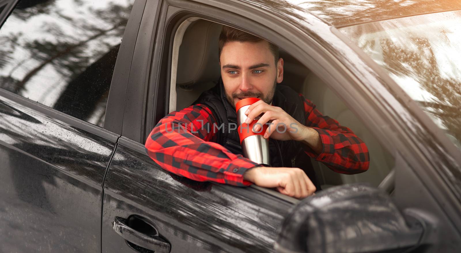 Young adult attractive Caucasian man sits at the wheel of his car sunny winter day. Wintertime road trip. Happy smiling hipster guy sitting in car and looking window. Drink hot beverage from thermos