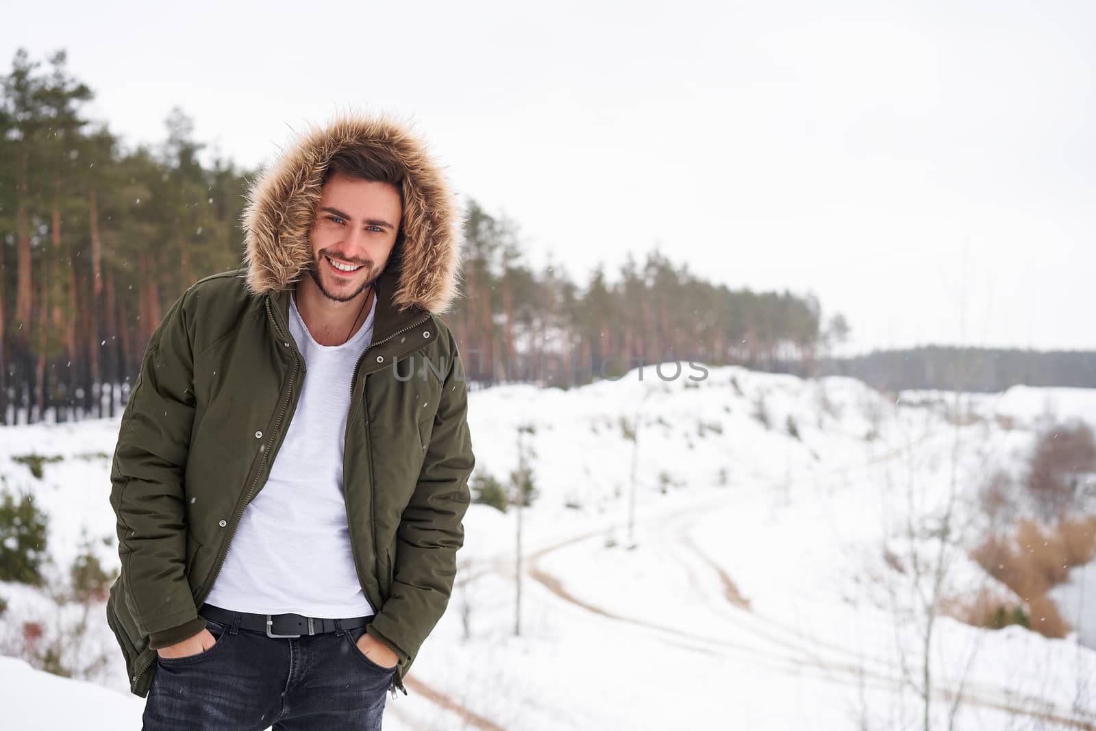 Handsome bearded young caucasian man standing outdoors fur hood in winter season forest. Attractive stylish european guy walking snowy christmas woodland Season holiday leisure