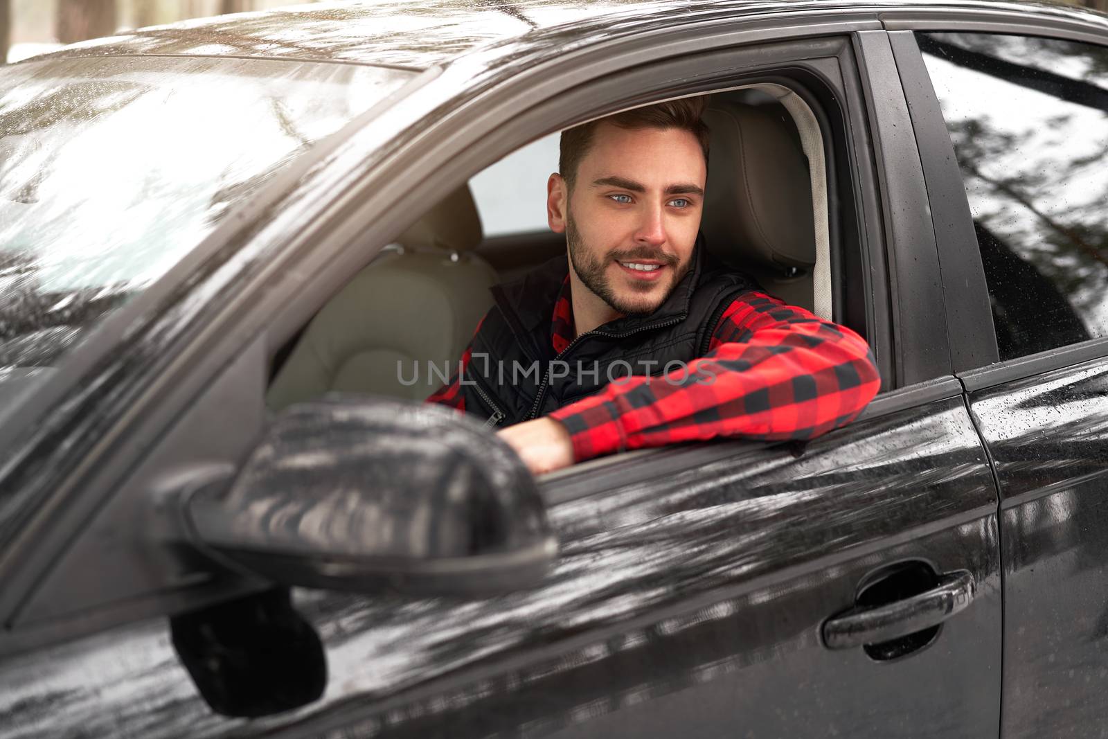 Young adult attractive Caucasian man sits at the wheel of his car sunny winter day. Wintertime road trip. Happy smiling hipster guy sitting in car and looking window. Portrait Positive driver