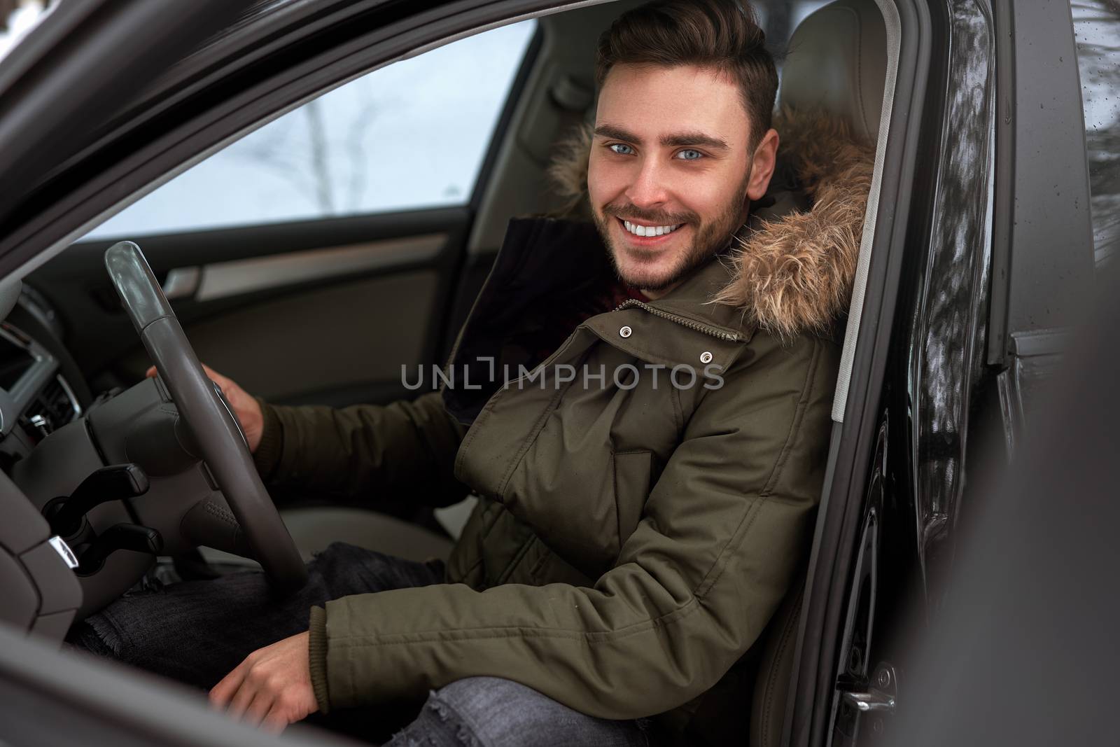 Young adult attractive Caucasian man sits at the wheel of his car sunny winter day. Wintertime road trip. Happy smiling hipster guy sitting in car and looking window. Portrait Positive driver