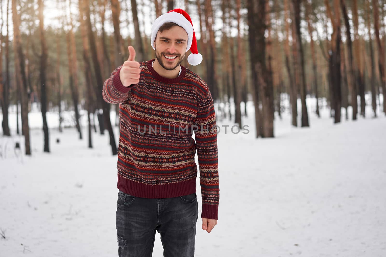 Handsome bearded young caucasian man standing outdoors sweater santa hat winter season forest. Attractive stylish european guy walking snowy christmas woodland Season holiday leisure Showing thumbs up