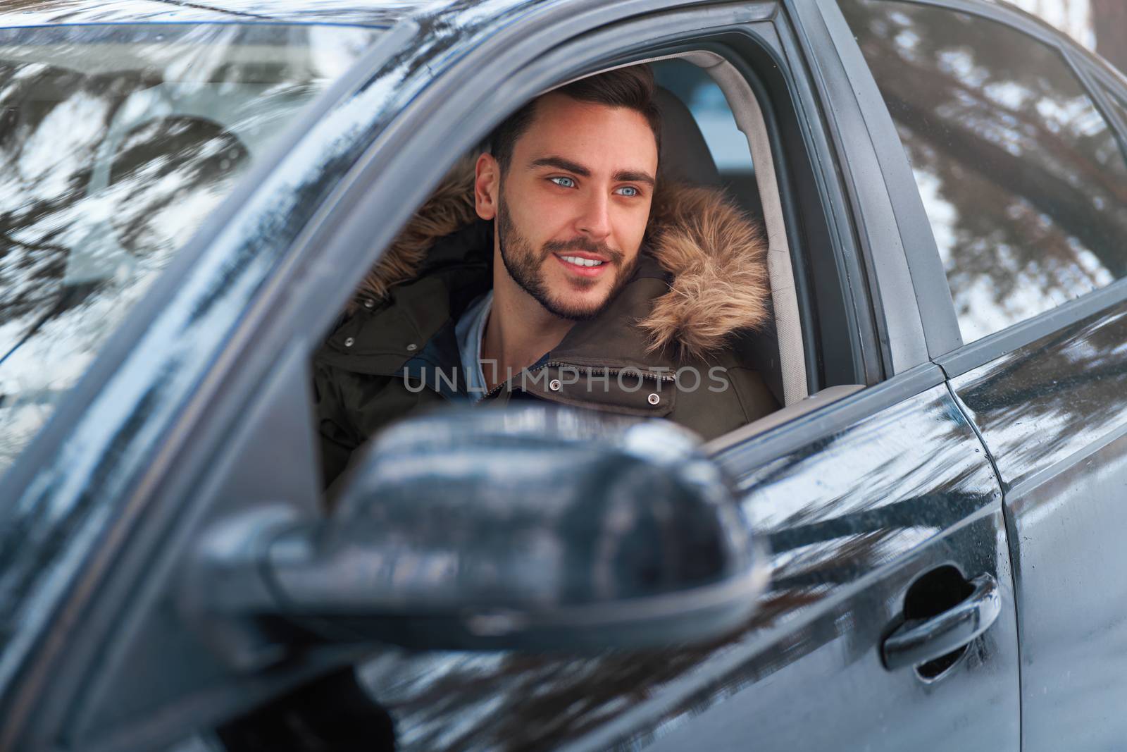 Young adult attractive Caucasian man sits at the wheel of his car sunny winter day. Wintertime road trip. Happy smiling hipster guy sitting in car and looking window. Portrait Positive driver