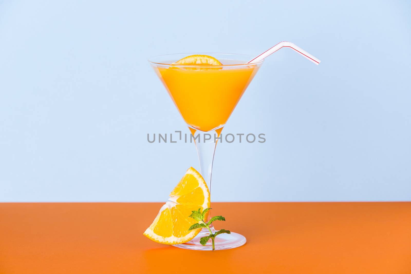 Fresh orange juice in a glass with a slice and a mint leafs at the base on an orange surface and a light blue background