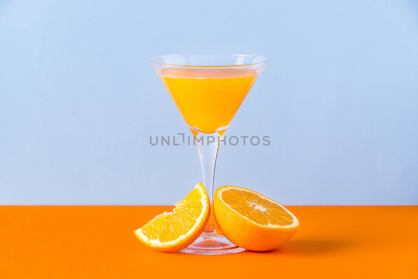 Fresh orange juice in a glass with a slice and a half at the base on an orange surface and a light blue background
