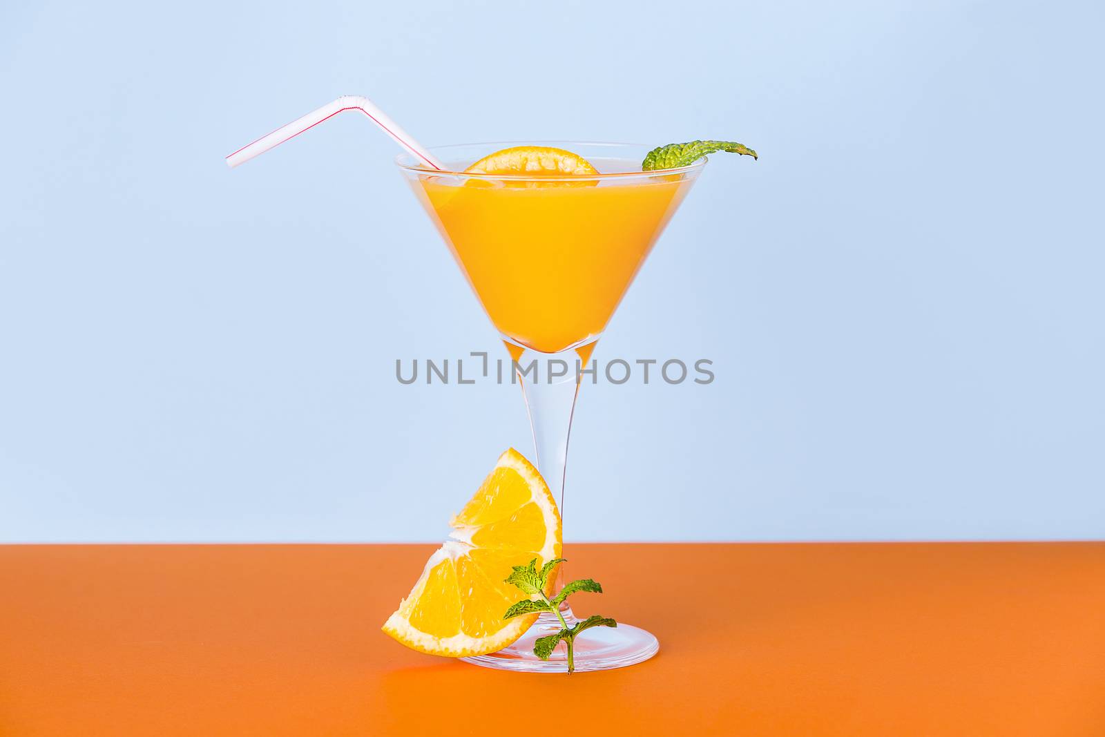 Fresh orange juice with a slice and a mint leaf at the base on an orange surface and a light blue background