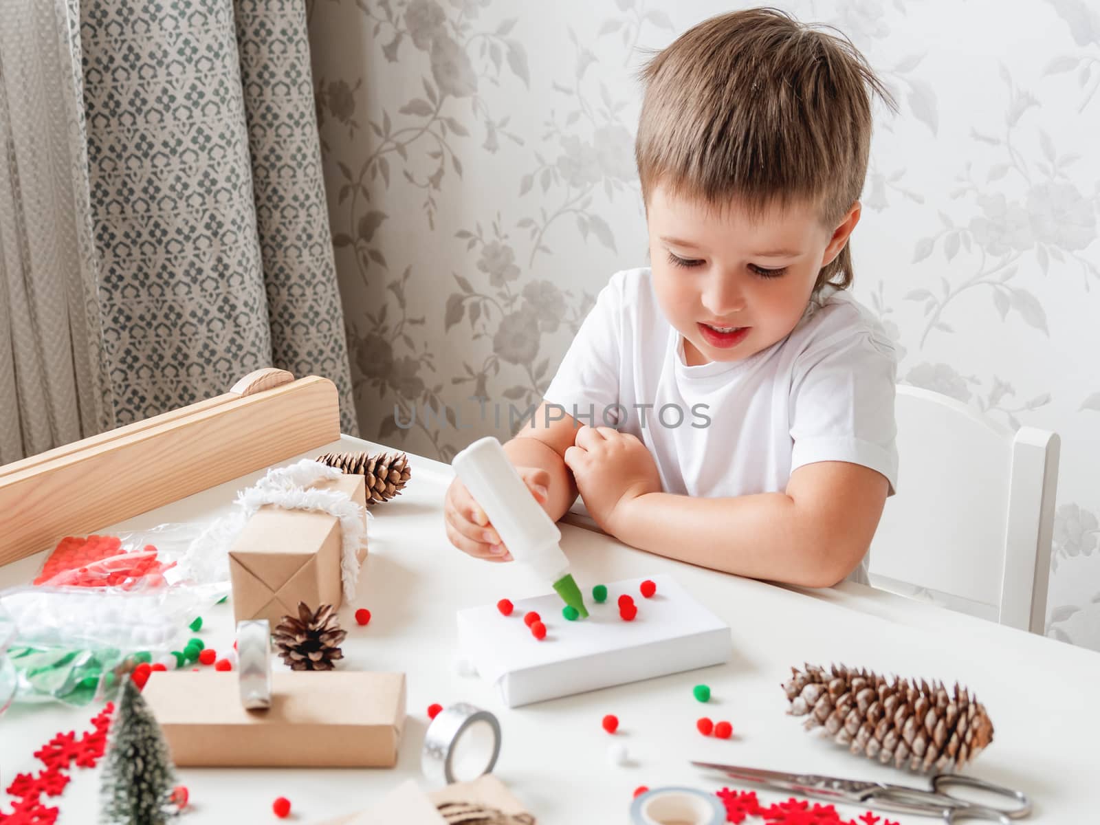 Child wrapped Christmas presents in craft paper with white fluffy pompons, feathers and fringe as decoration. White table with handmade New Year gifts.