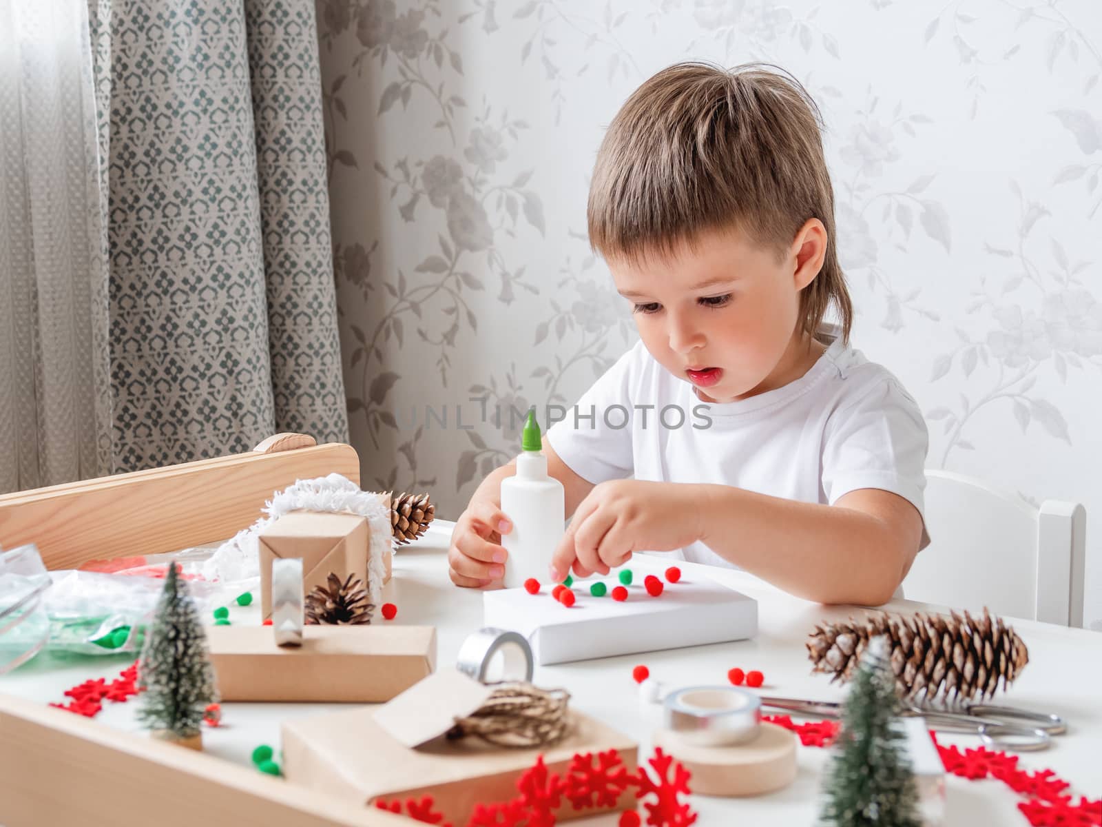 Child wrapped Christmas presents in craft paper with white fluffy pompons, feathers and fringe as decoration. White table with handmade New Year gifts.