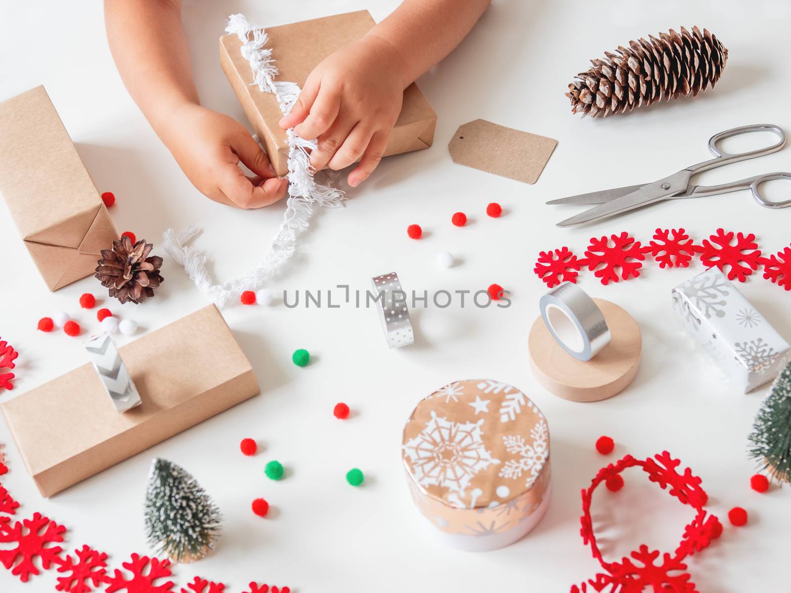 Kid wraps handmade Christmas presents in craft paper with colorful pompons and snowflake ribbons. Child prepares gifts for New Year celebration. Peaceful leisure activity before winter holidays.