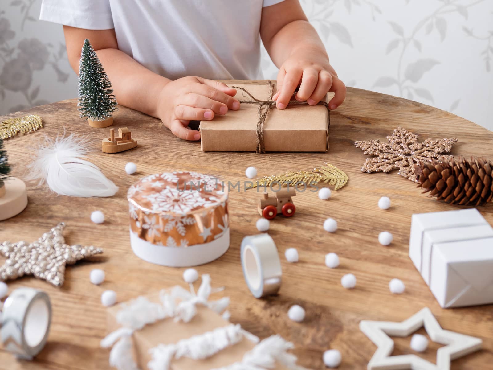 Child wrapped Christmas presents in craft paper with white fluffy pompons, feathers and fringe as decoration. Wooden table with hand made New Year gifts.