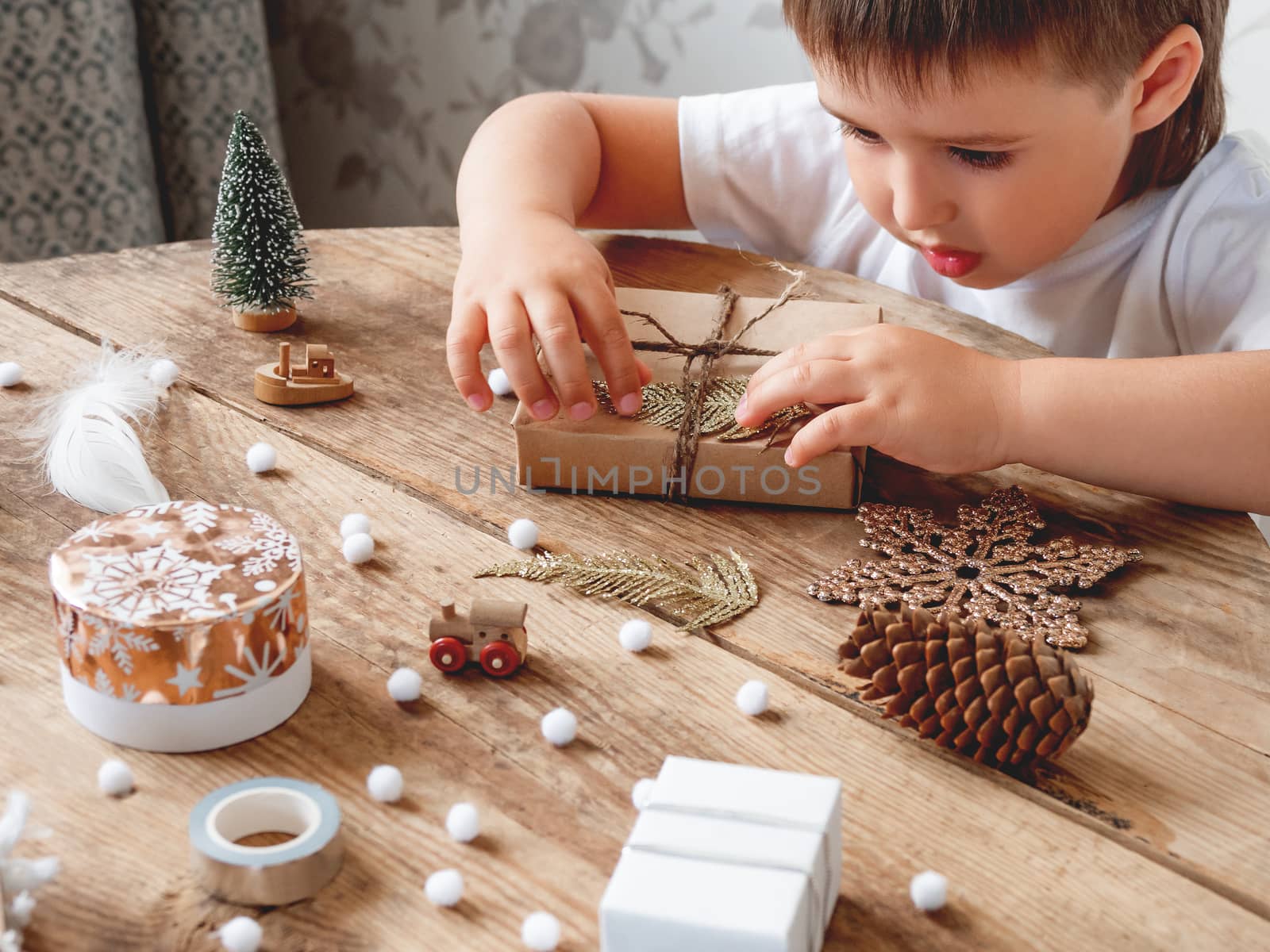 Child wrapped Christmas presents in craft paper with white fluffy pompons, feathers and fringe as decoration. Wooden table with hand made New Year gifts.