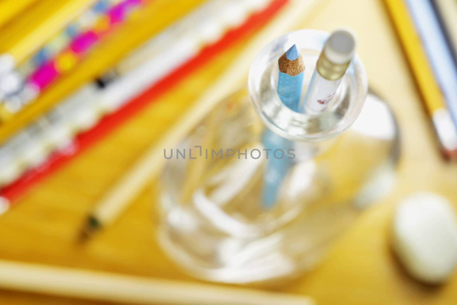 Wooden work desk with colored pencils , in the foreground pencil box with blue pencil and pencil eraser , a glass bottle is used as box  ,macro photography